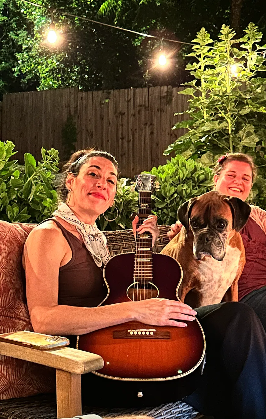 Photo of two people sitting outside, one holding a guitar, with Otis the dog between them